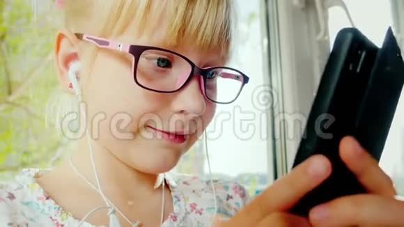 有趣的女孩微笑着用电话她戴着玫瑰色眼镜和白色耳机快乐孩子的肖像视频的预览图