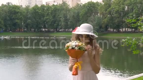 美丽的年轻女孩穿着一件浅色的连衣裙和一顶白色的帽子在公园里散步视频的预览图