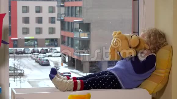 可爱的小女孩躺在靠窗户的散热器上带着一只泰迪熊冬天下雪视频的预览图