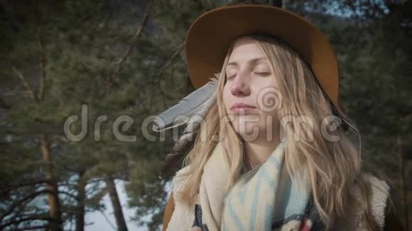 年轻的女旅行者看着太阳日落或黎明戴上帽子和背包在一棵松树的背景下视频的预览图