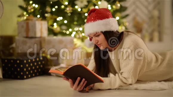 快乐惊讶的女人戴着圣诞帽长发躺在地板上享受阅读微笑的特写肖像视频的预览图