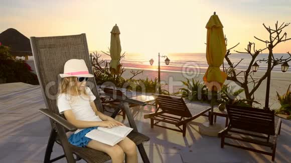 年轻的白种人女孩坐在扶手椅上看书海上日落长椅子和雨伞有相同的剪辑视频的预览图