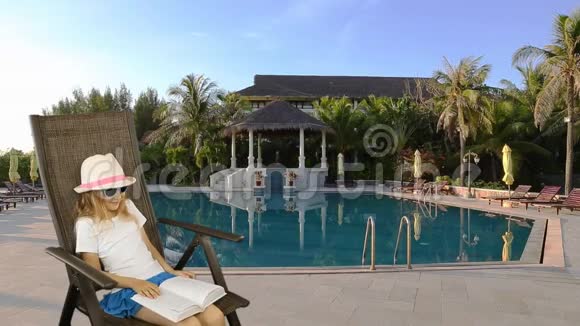 年轻的白种人女孩坐在扶手椅上看书游泳池和背景棕榈树同样的夹子视频的预览图