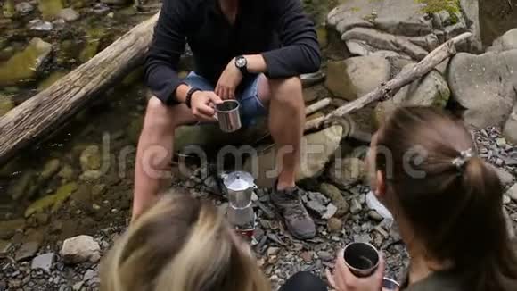 一个男人和两个女孩背包客在溪边露营时喝咖啡视频的预览图