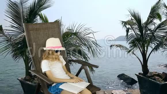 年轻的白种人女孩坐在扶手椅上看书海岸地平线棕榈树的背景同样的夹子视频的预览图