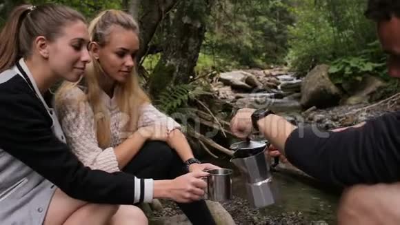 一个男人在溪边的营地里用杯子把咖啡倒给女孩们视频的预览图