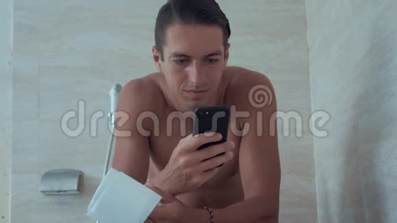 年轻人用智能手机坐在家里的厕所里坐在马桶里拿电话的人视频的预览图