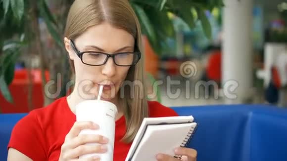 聪明的口渴学生戴着眼镜坐在咖啡馆里喝着纸杯里的可乐写在笔记本上视频的预览图
