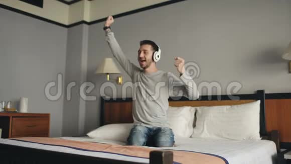 在酒店房间里一个年轻快乐的男人用无线耳机听音乐在床上跳舞视频的预览图