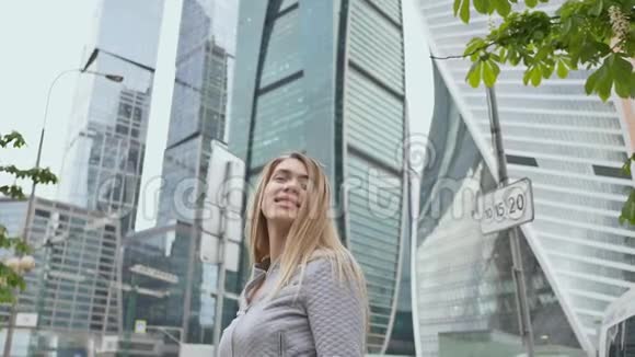 年轻快乐的金发女孩在高楼大厦和摩天大楼的背景下竖起大拇指视频的预览图