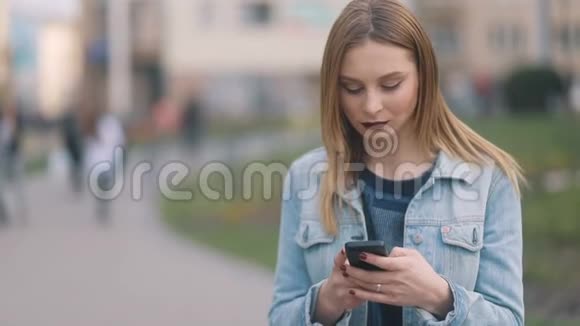 性感的金发女郎深红的嘴唇穿着蓝色牛仔布的女孩以及通过智能手机进行严格的模糊交流城市视频的预览图