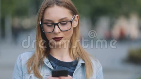 欧洲温柔的极客女孩深红色的嘴唇在蓝色牛仔法庭和严格的模糊使用她的智能手机在城市视频的预览图
