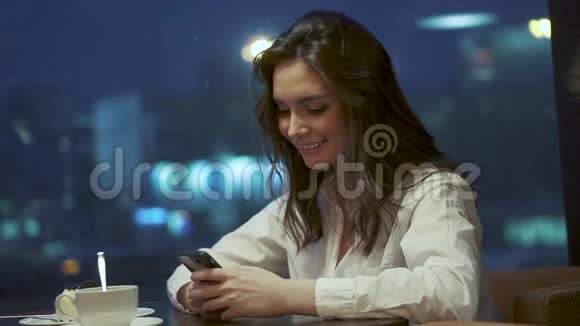 年轻女孩正在智能手机上发短信坐在夜晚灯光背景下的咖啡馆里视频的预览图