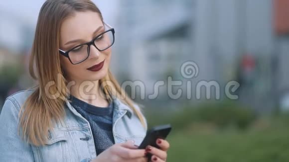 在街头背景下戴着眼镜的漂亮年轻女孩使用她的移动智能手机的肖像慢吞吞视频的预览图