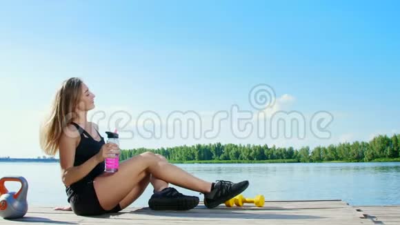 美丽的运动的金发女孩女人在户外训练后休息喝水湖泊河流蓝天和森林视频的预览图