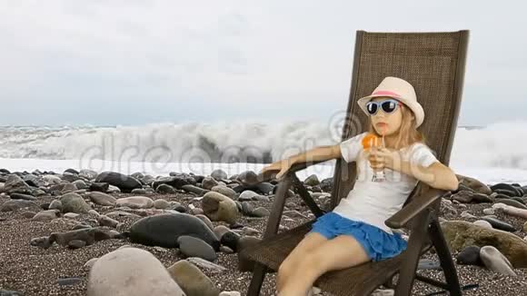 年轻的白种人女孩坐在扶手椅上用吸管喝橙汁鸡尾酒海岸线棕色视频的预览图