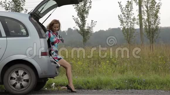 一个金发女孩坐在她的破车后备箱里在一条乡村道路上闪烁着紧急信号视频的预览图