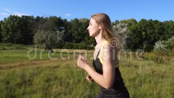 美丽的高高的运动的金发女郎穿着上衣和短裤的女孩在绿色的草地小径上慢跑视频的预览图