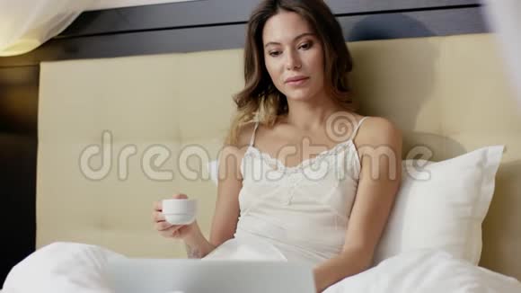 美丽的女人在床上一边喝咖啡一边用手提电脑看新闻视频的预览图