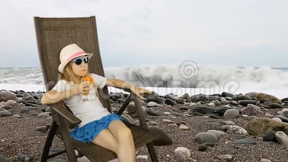 年轻的白种人女孩坐在扶手椅上用吸管喝橙汁鸡尾酒海岸线棕色视频的预览图