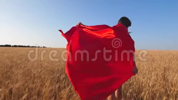 女孩超级英雄穿着红色斗篷在蓝天上跑过田野慢动作视频的预览图