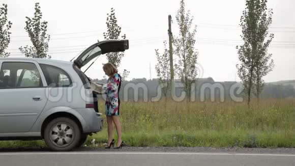 一个金发女孩在一条乡间小路上的一辆开着后备箱的破车旁站着读着说明书视频的预览图