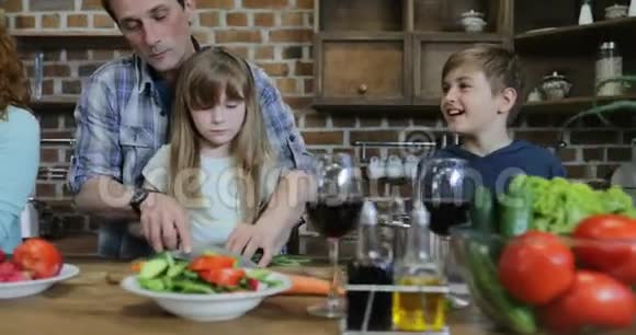 父亲和女儿一起切菜做饭在厨房和儿子说话而母亲使用平板电脑数字视频的预览图