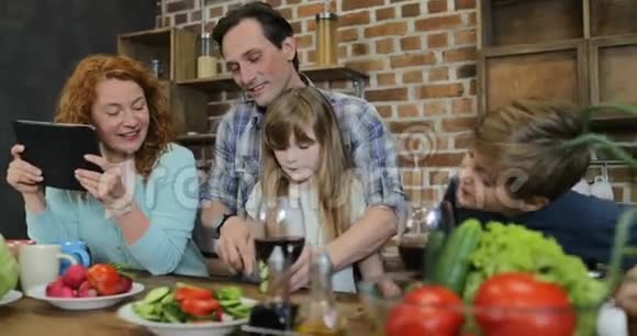 父亲和女儿一起切菜做饭在厨房和儿子说话而母亲使用平板电脑数字视频的预览图