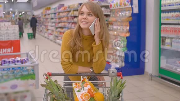 漂亮的年轻女子在超市打电话她推着一辆购物车视频的预览图