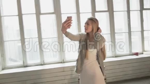 快乐美丽的年轻女人在靠窗的地方自拍带着智能手机笑的金发美女给自己拍照视频的预览图