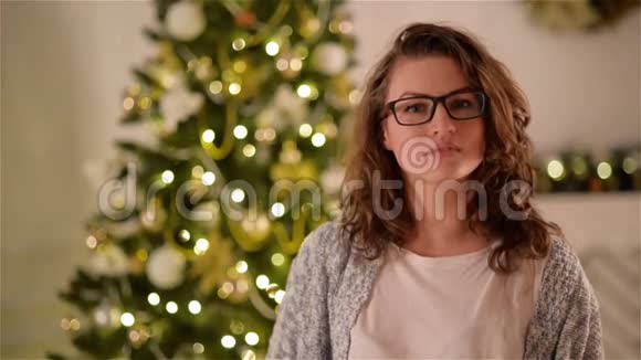 美丽的女孩长着卷发和眼镜看着相机和微笑的圣诞树背景特写视频的预览图