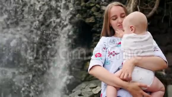 母亲带着孩子微笑拥抱婴儿的儿子关于瀑布亲吻和与他交谈视频的预览图