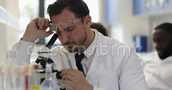 在实验室穿着白色外套和防护眼镜的男性科学家用显微镜观察试管视频的预览图