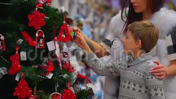 快乐的母亲和她的小儿子正在商场里观察圣诞树上的装饰特写侧视视频的预览图