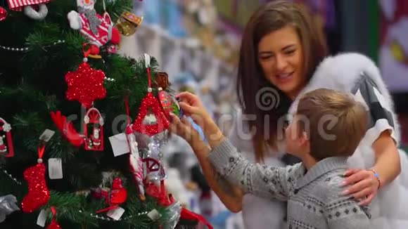 这位活泼的母亲和她的儿子在圣诞树上观察可爱的装饰物的特写侧画视频的预览图