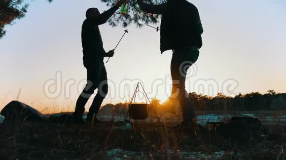 快乐的两个旅游朋友在日落时坐在森林的篝火旁取暖团体旅行者野餐视频的预览图