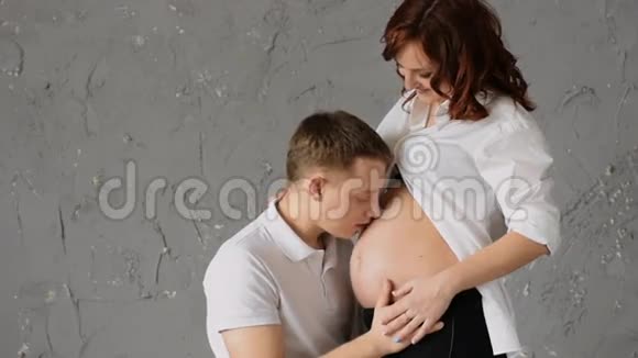 美丽的年轻人亲吻他怀孕妻子的腹部年轻的妻子抚摸他的头看着他视频的预览图