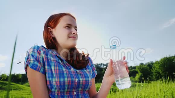 在风景如画的地方野餐时一个青少年从瓶子里喝干净的水美丽的绿色草地和蓝天视频的预览图