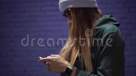 一个时髦的时髦女孩戴着时髦的眼镜戴着一顶帽子智能手机上的应用程序就在砖紫墙附近视频的预览图