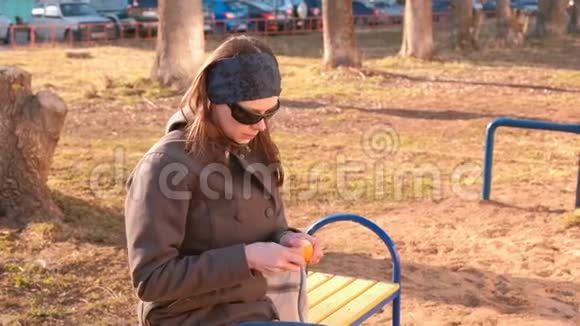 年轻的黑发女人戴着太阳镜剥着橘子坐在公园的长凳上视频的预览图