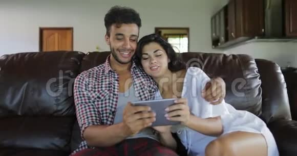 夫妻拥抱坐在教练用平板电脑在客厅年轻人和女人在网上浏览现代视频的预览图