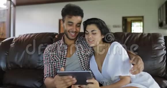 夫妻拥抱坐在教练用平板电脑在客厅年轻人和女人在网上浏览现代视频的预览图