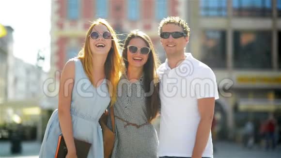 三个微笑的朋友在时尚太阳镜正在看相机同时在城市背景下拍摄户外视频的预览图