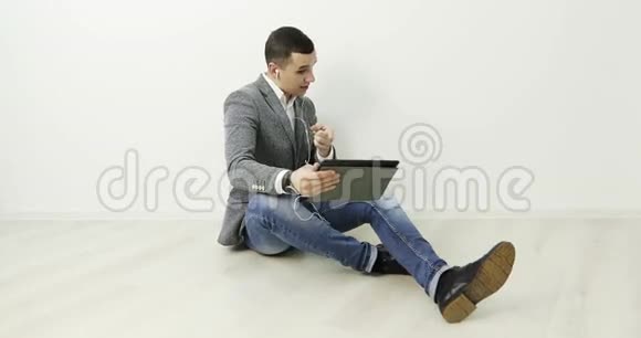 4K一个年轻时尚的家伙坐在办公室的地板上用耳机听音乐视频的预览图