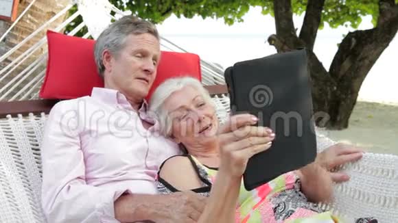 使用数码平板电脑在沙滩吊床放松老年夫妇视频的预览图