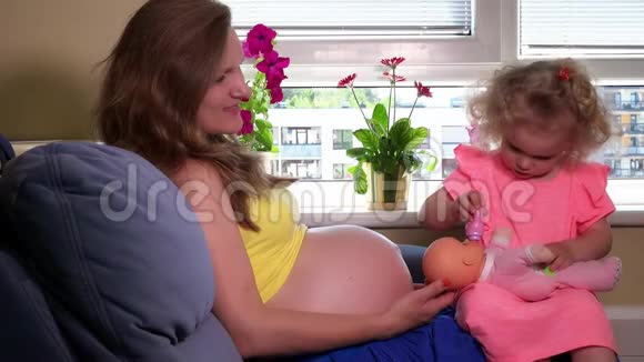 可爱的小女孩喂玩具宝宝瓶子坐在怀孕的母亲膝盖上视频的预览图