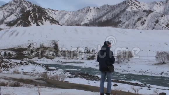 一位游客站在河岸边环顾四周冬天的群山视频的预览图