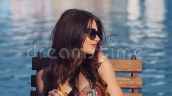 靠近美丽的年轻女子穿着比基尼喝鸡尾酒坐在游泳池旁晒日光浴视频的预览图