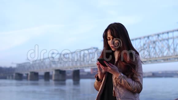 在桥边附近的河边使用智能手机的年轻女子视频的预览图