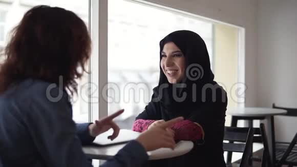 两位女性朋友在咖啡馆里交谈身穿黑头巾的年轻穆斯林妇女正在与她的女性白种人朋友交谈视频的预览图